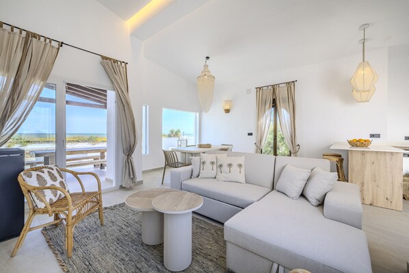 Uma sala de estar moderna com um sofá, uma mesa de centro redonda, uma poltrona de vime e vista para a praia.