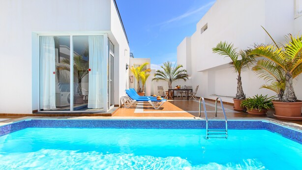 Piscina com borda em azulejos azuis, entrada por porta de vidro e área de estar com cadeiras e plantas em vasos.