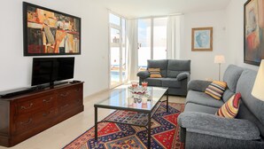 Uma sala de estar com televisão de tela plana, mesa de centro de vidro, sofá cinza e um tapete estampado.