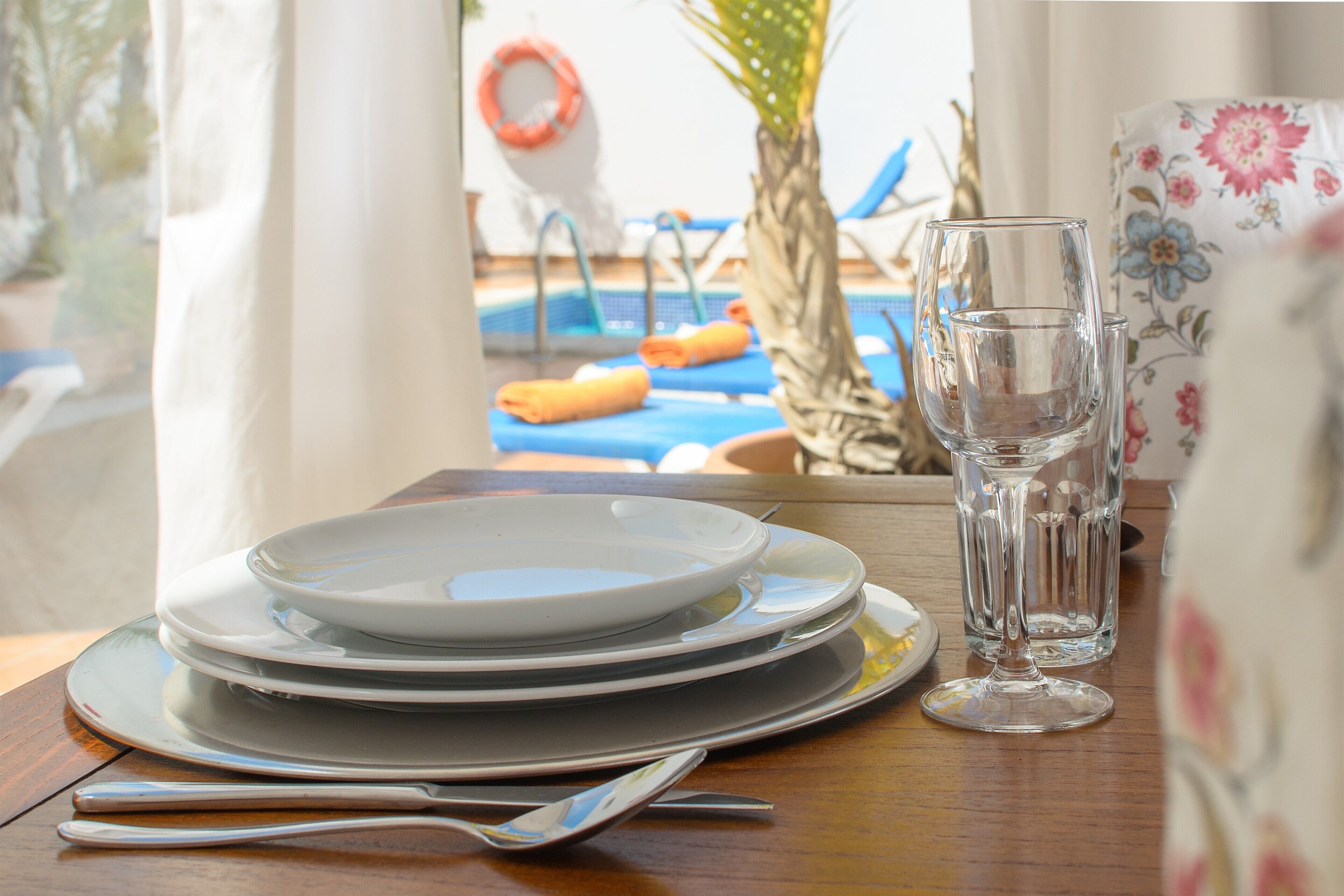 A table set with plates, a glass, and cutlery, with a pool and lounge chairs visible through a window.