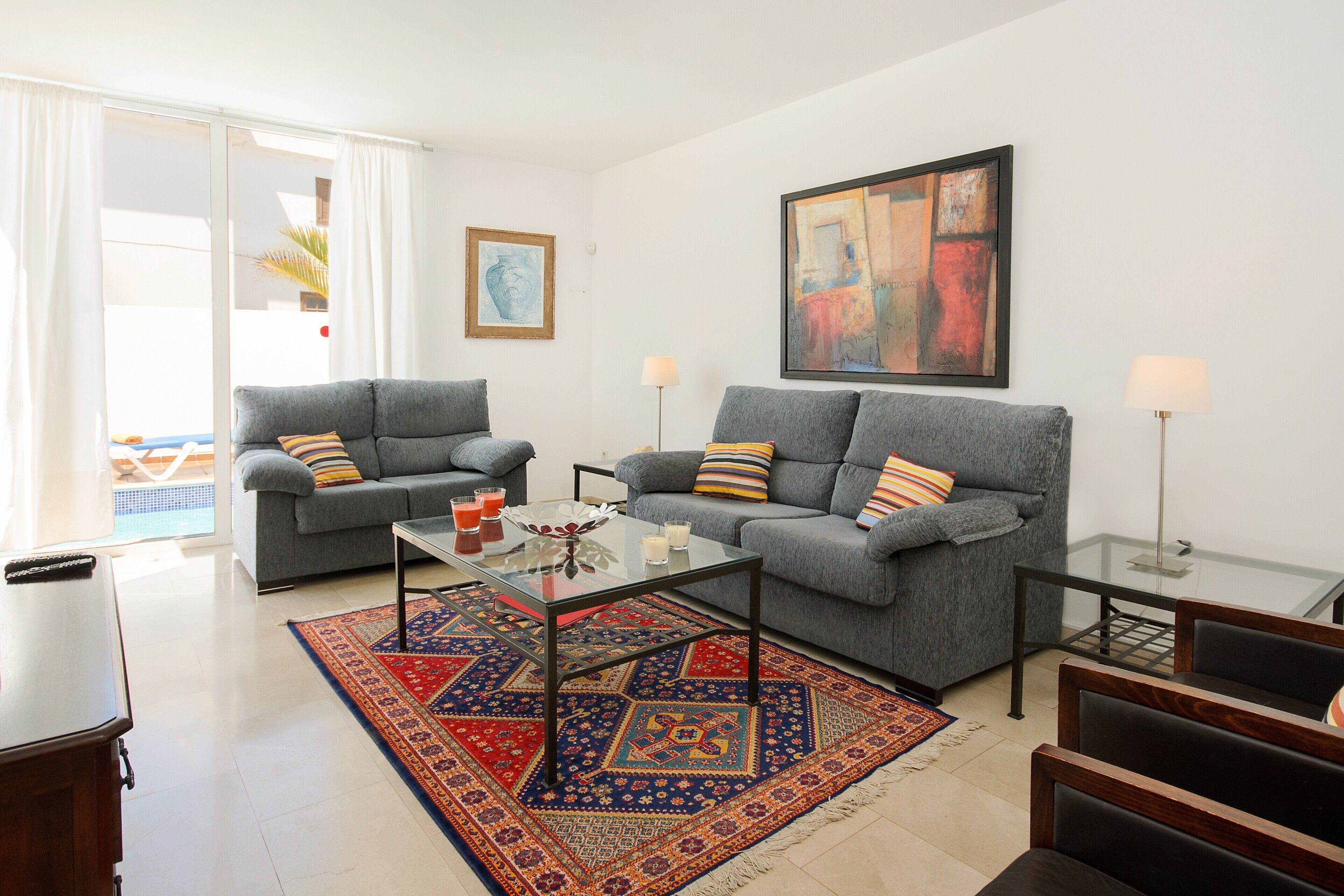 Uma sala de estar com dois sofás cinzas, uma mesa de centro de vidro, um tapete estampado e um quadro na parede.