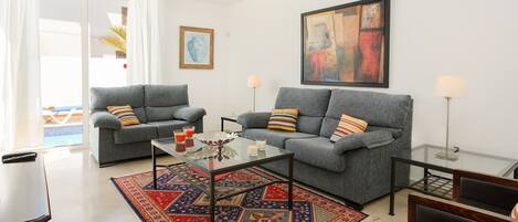 Uma sala de estar com dois sofás cinzas, uma mesa de centro de vidro, um tapete estampado e um quadro na parede.