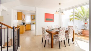 Uma sala de jantar com mesa de madeira e cadeiras florais, uma cozinha ao fundo e uma janela grande com vista para uma palmeira.