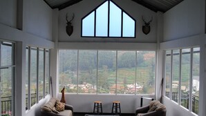 A room with a large window offering a view of a green landscape, two chairs, a small table, and two wall-mounted animal heads. - Misty Mountain Villas (Nuwara Eliya)