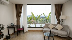 Quarto de hotel com cama, sofá, escrivaninha com laptop, mesa pequena com garrafa e copos, e vista para vegetação e montanhas através de amplas janelas.