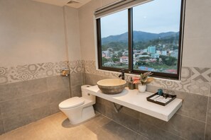 A modern bathroom with a large window, a white toilet, a beige sink with a large bowl, and a small shelf with toiletries. - Max Boutique Hotel (Thuc Phan)