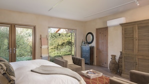 A bedroom with a large bed, a chair, a small table, and a wardrobe. There is a window with a view of greenery outside.