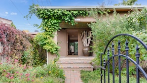 A garden entrance with a brick pathway leading to a door, surrounded by lush greenery and a large vase.