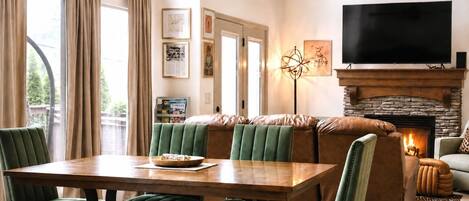 A cozy living room with a fireplace, a dining area with a wooden table and green upholstered chairs, and a large flat-screen TV mounted above the fireplace.