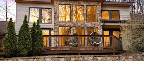 A modern two-story house with a stone facade, large windows, and a wooden deck with outdoor seating.