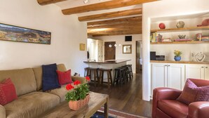 Uma sala de estar aconchegante com um sofá, uma mesa de centro com uma planta e uma poltrona de couro vermelha. Há uma área de cozinha com banquetas e uma estante com diversos objetos.