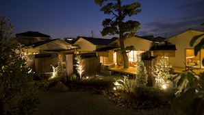A residential area with houses at dusk, featuring a well-lit patio area with a dining table and chairs, surrounded by trees and a small garden. - Migiwaya (Yaizu)