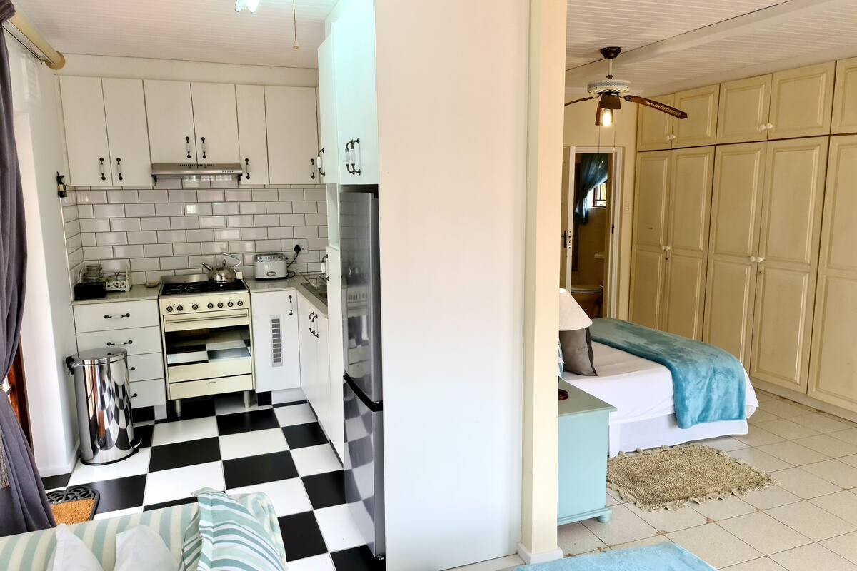 A compact kitchen with white cabinets, a black and white checkered floor, a bed with a blue blanket, and a small table with a lamp.