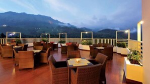 An outdoor seating area with wicker furniture and tables, overlooking a mountainous landscape. - Gokulam Park Munnar (Devikolam)