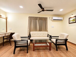 A room with a ceiling fan, an air conditioning unit, a desk with a chair, a sofa, and a coffee table. - Hotel Arya Palace (Bhubaneshwar)