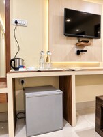 Habitación de hotel con televisor de pantalla plana montado en la pared, un refrigerador pequeño, cafetera y una mesa con hervidor, vasos y un plato.
