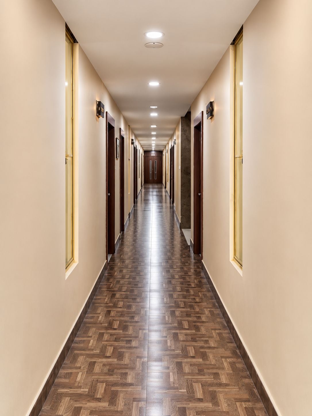 Un lungo e stretto corridoio di un albergo, con porte su entrambi i lati, pavimento in legno decorato e illuminazione a incasso.