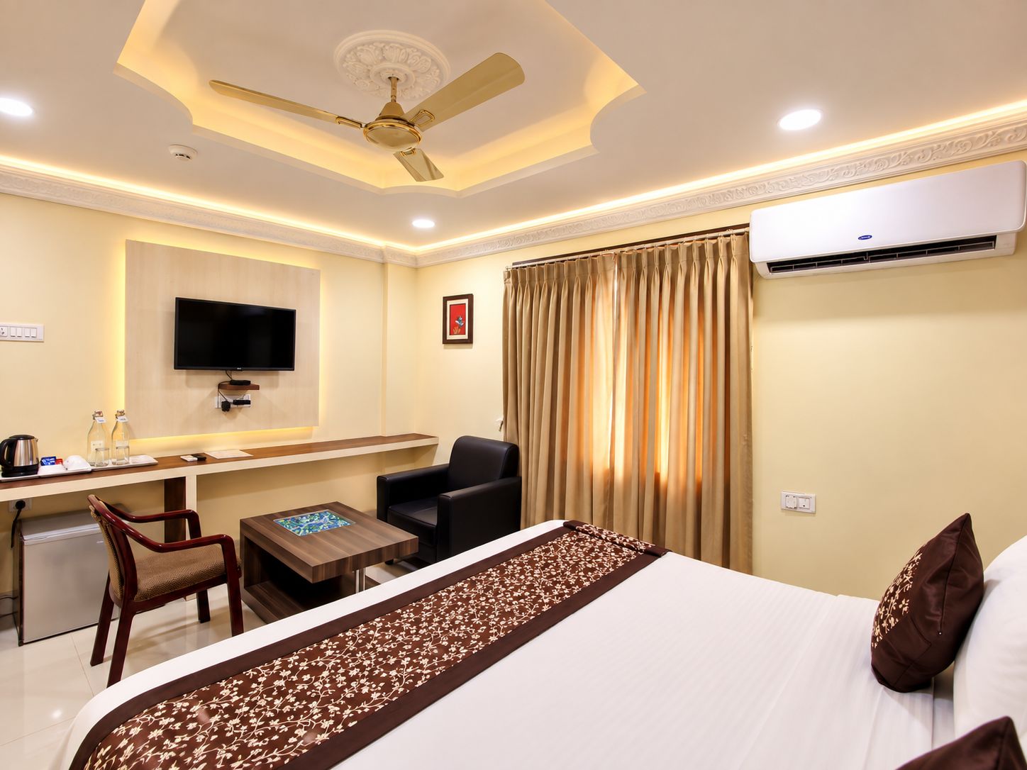 A hotel room with a bed, a desk with a chair, a television, an air conditioning unit, and a ceiling fan.