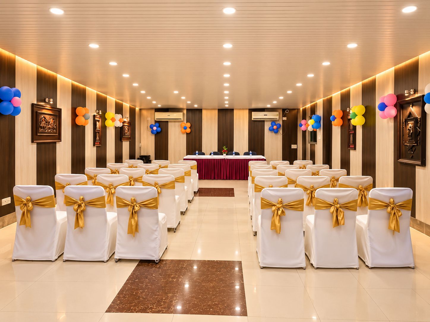 Ein Konferenzraum mit Reihen weißer Stühle mit goldenen Bändern, einem langen Tisch mit roter Tischdecke und bunten Ballons an den Wänden.