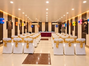 Un salón de conferencias con filas de sillas blancas con cintas doradas, una mesa larga con mantel rojo y globos de colores en las paredes.