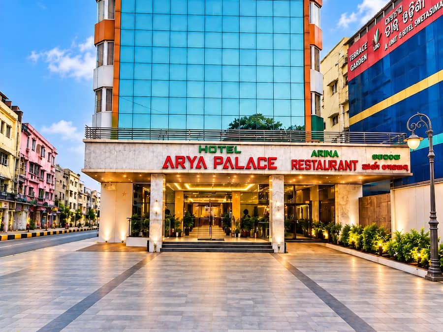 Un hotel moderno con fachada de vidrio, llamado 'Arya Palace', que presenta una entrada destacada y el letrero visible de un restaurante.