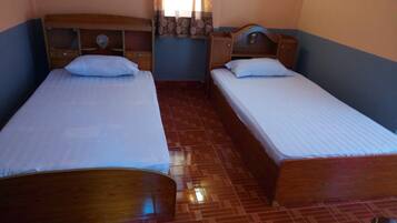 A room with two wooden beds with white linens, a wooden nightstand with a mirror, and a patterned curtain.