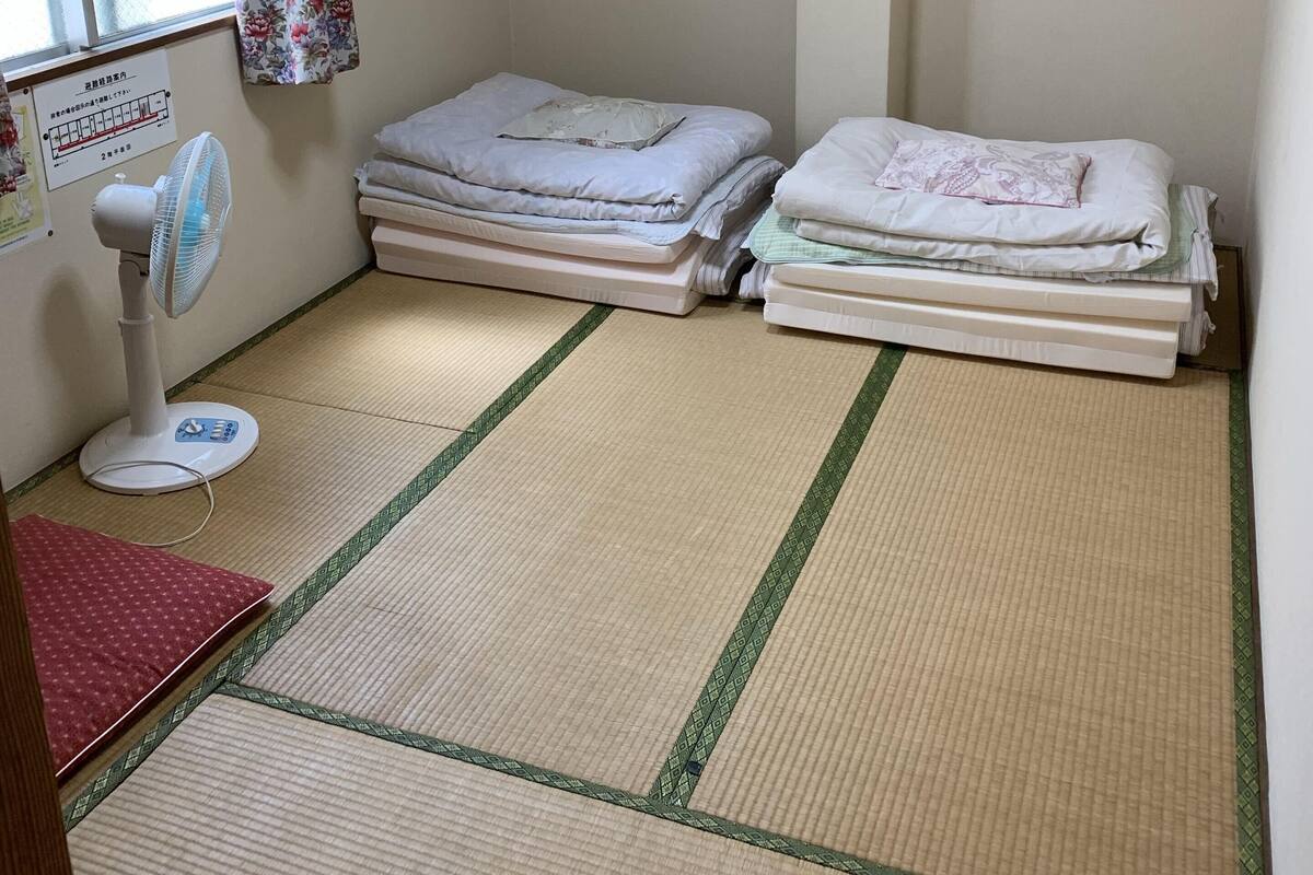 Een kamer met tatami vloerbedekking, twee bedden met opgemaakt beddengoed, een staande ventilator en een raam met bloemen gordijnen.