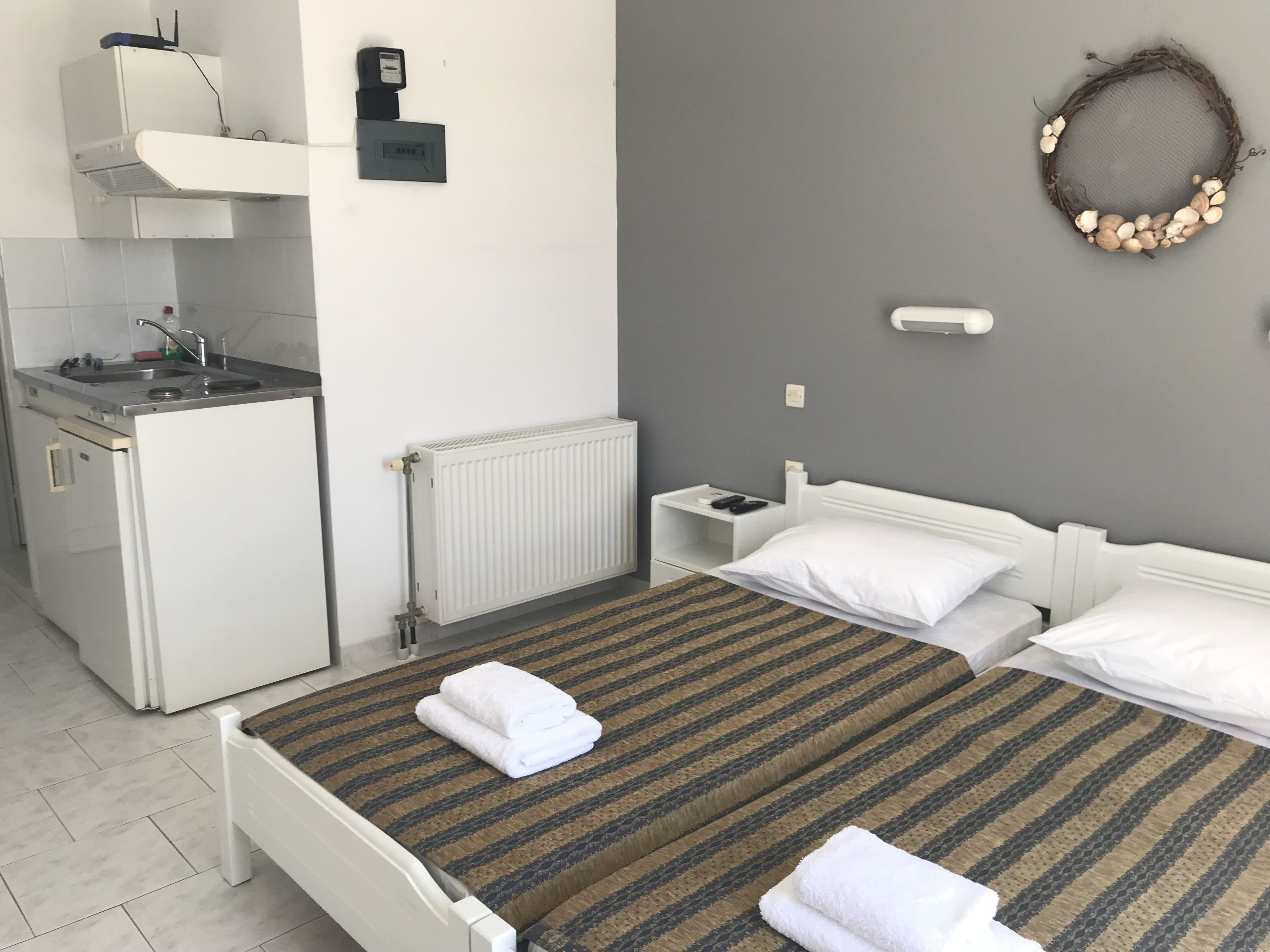 A compact hotel room with a kitchenette, a bed with two pillows, a small bedside table, and a wall-mounted light fixture.