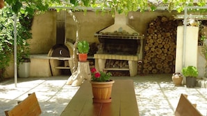 An outdoor patio with a wooden table and benches, a pizza oven, and a stack of firewood. - Villa Katerina (Syros)