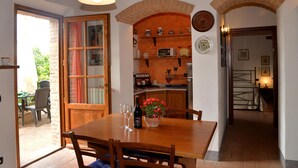 A dining area with a wooden table, chairs, and a bottle of wine. There is a kitchen area with shelves and a refrigerator in the background.