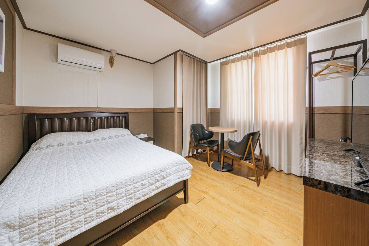 Una habitación de hotel con cama, una mesita, dos sillas, un espejo y una ventana con cortinas.