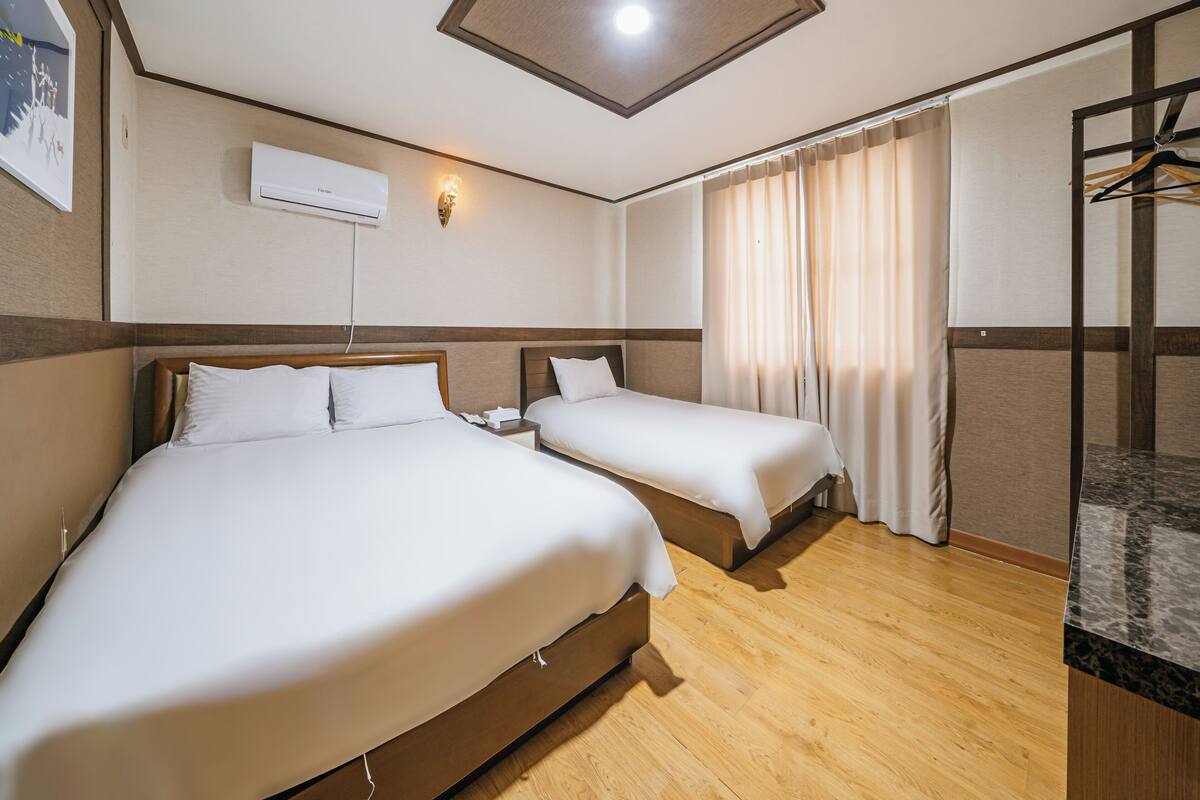 Habitación de hotel con dos camas, suelo de madera, aire acondicionado instalado en la pared y baño con ducha y barra para toallas.
