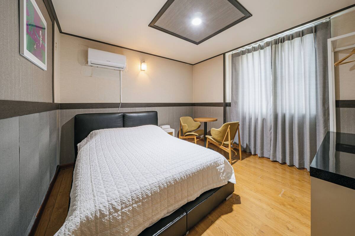 Habitación de hotel con una cama grande, una mesita pequeña, dos sillas, una lámpara de techo y una ventana con cortinas.
