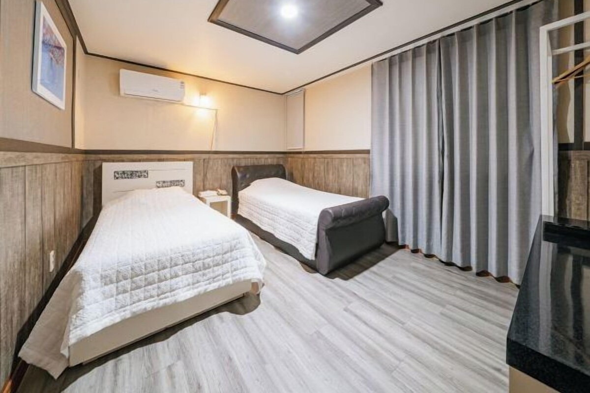 A hotel room with two beds, a wooden floor, a ceiling light, and a window with curtains.