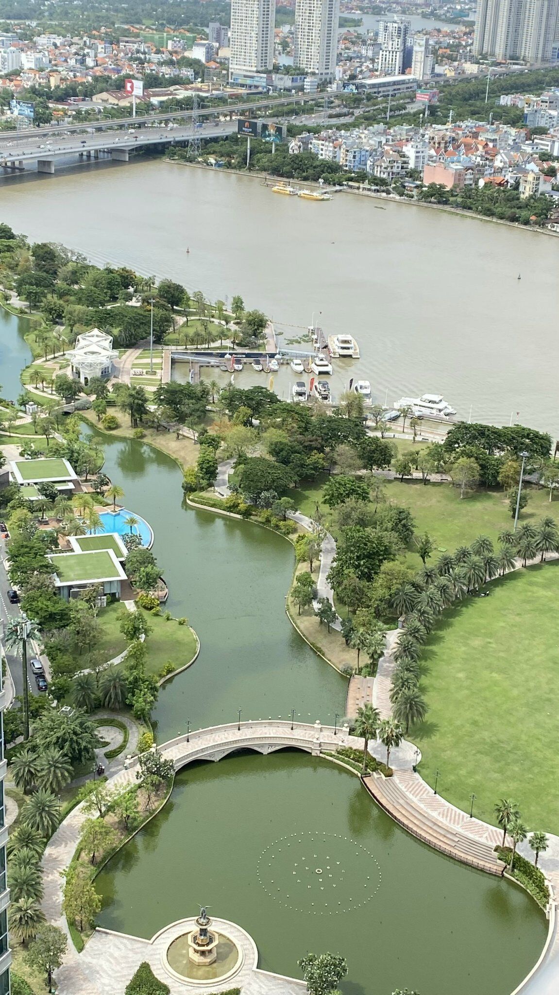A large park with a central pond, surrounded by greenery and a bridge.