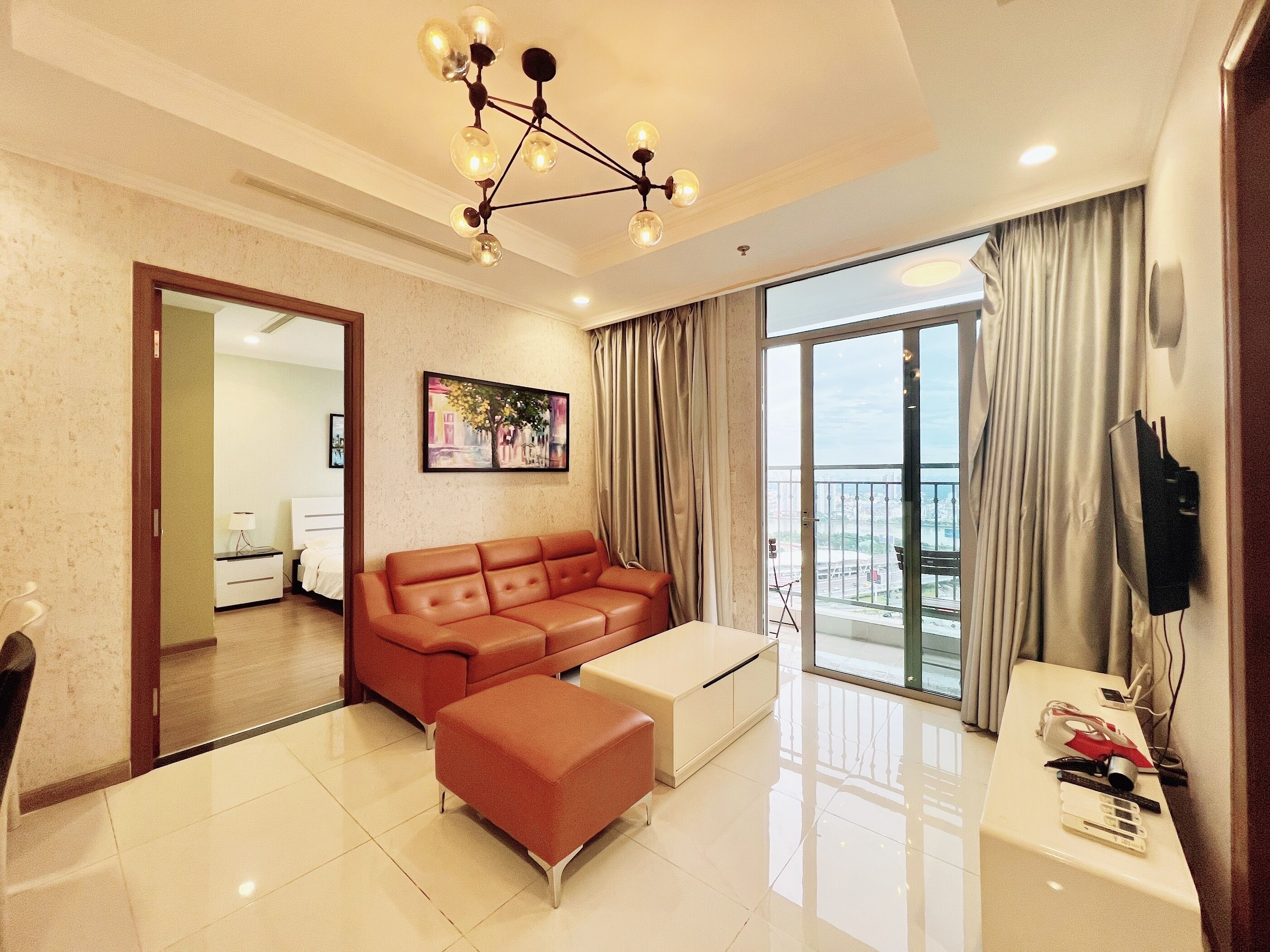 A modern living room with a sofa, ottoman, and a coffee table. There is a painting on the wall, a ceiling light fixture, and a balcony with curtains.