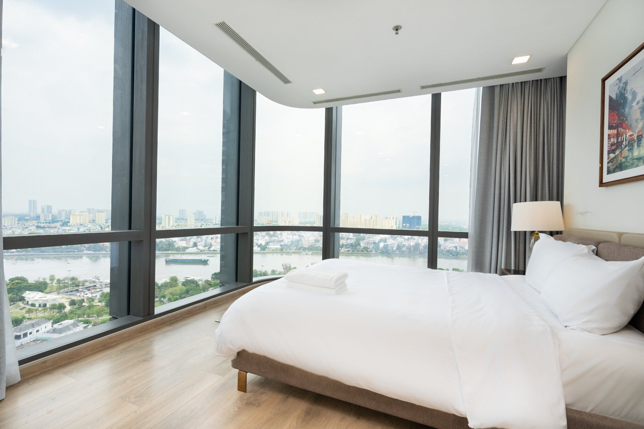 A hotel room with a large bed, a view of a cityscape through floor-to-ceiling windows, and a painting on the wall.