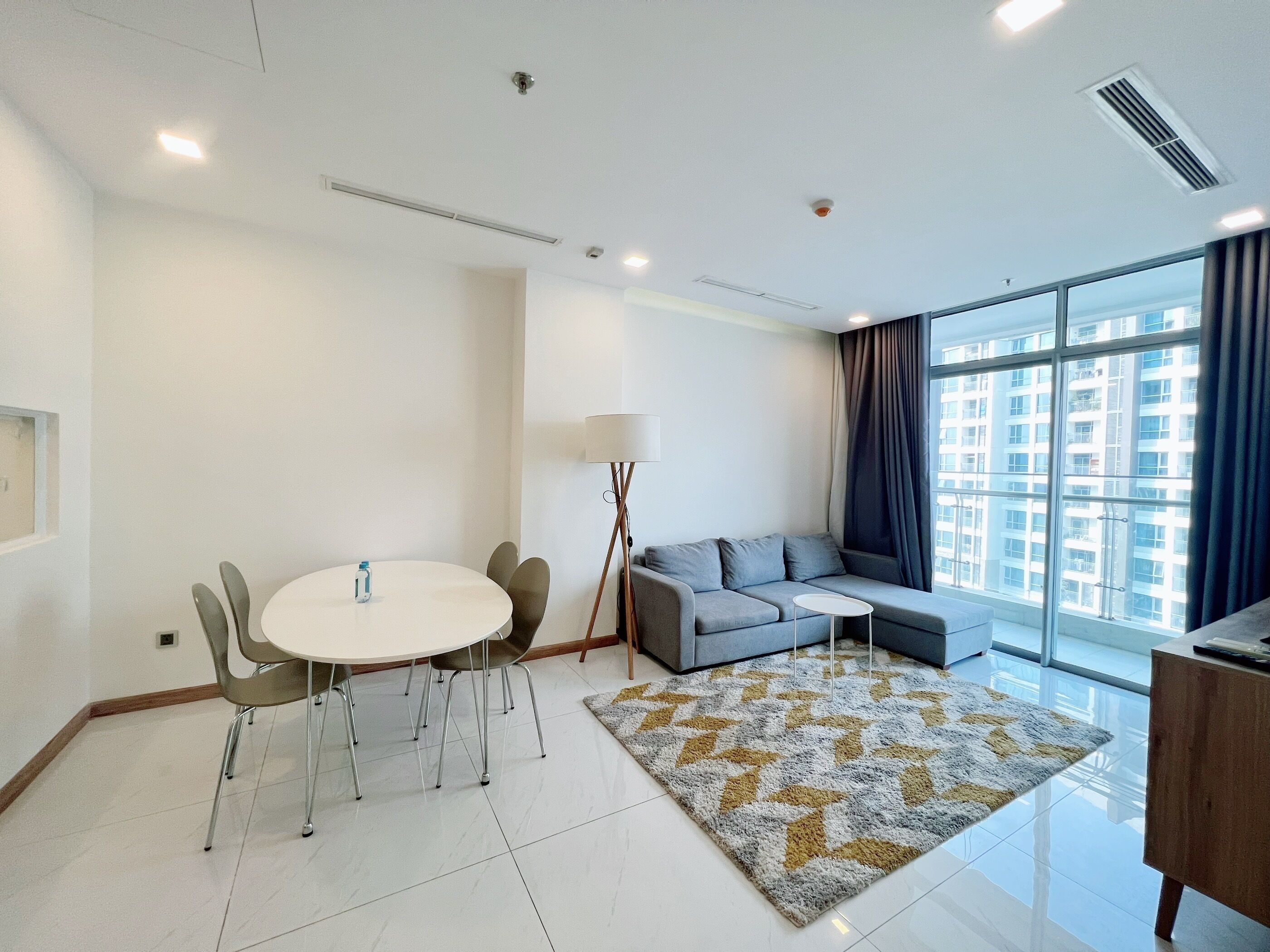 A modern living room with a round dining table, grey sofa, and a large window with curtains.