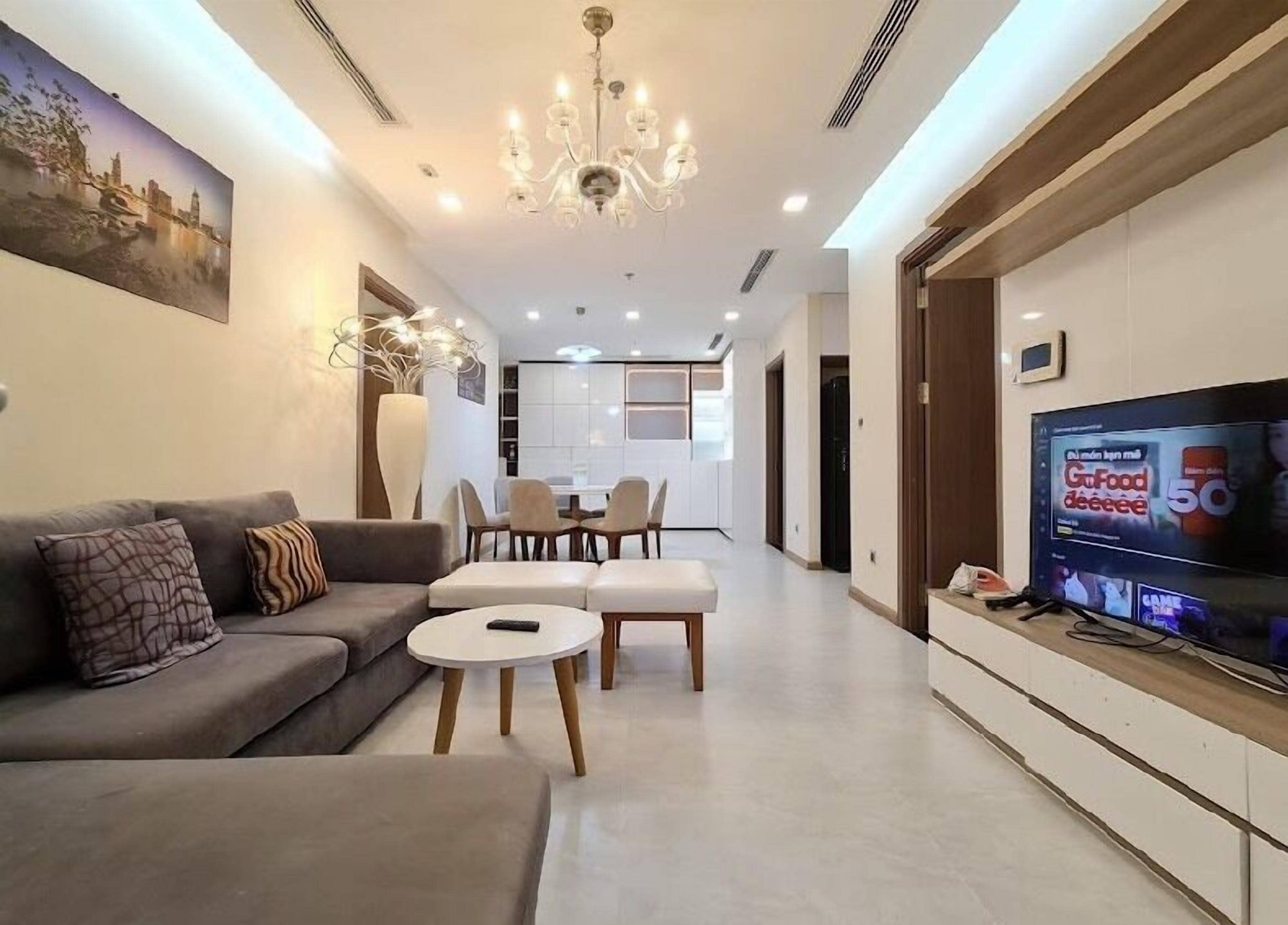 A modern living room with a sofa, coffee table, and TV. There is a dining area with chairs and a table in the background.