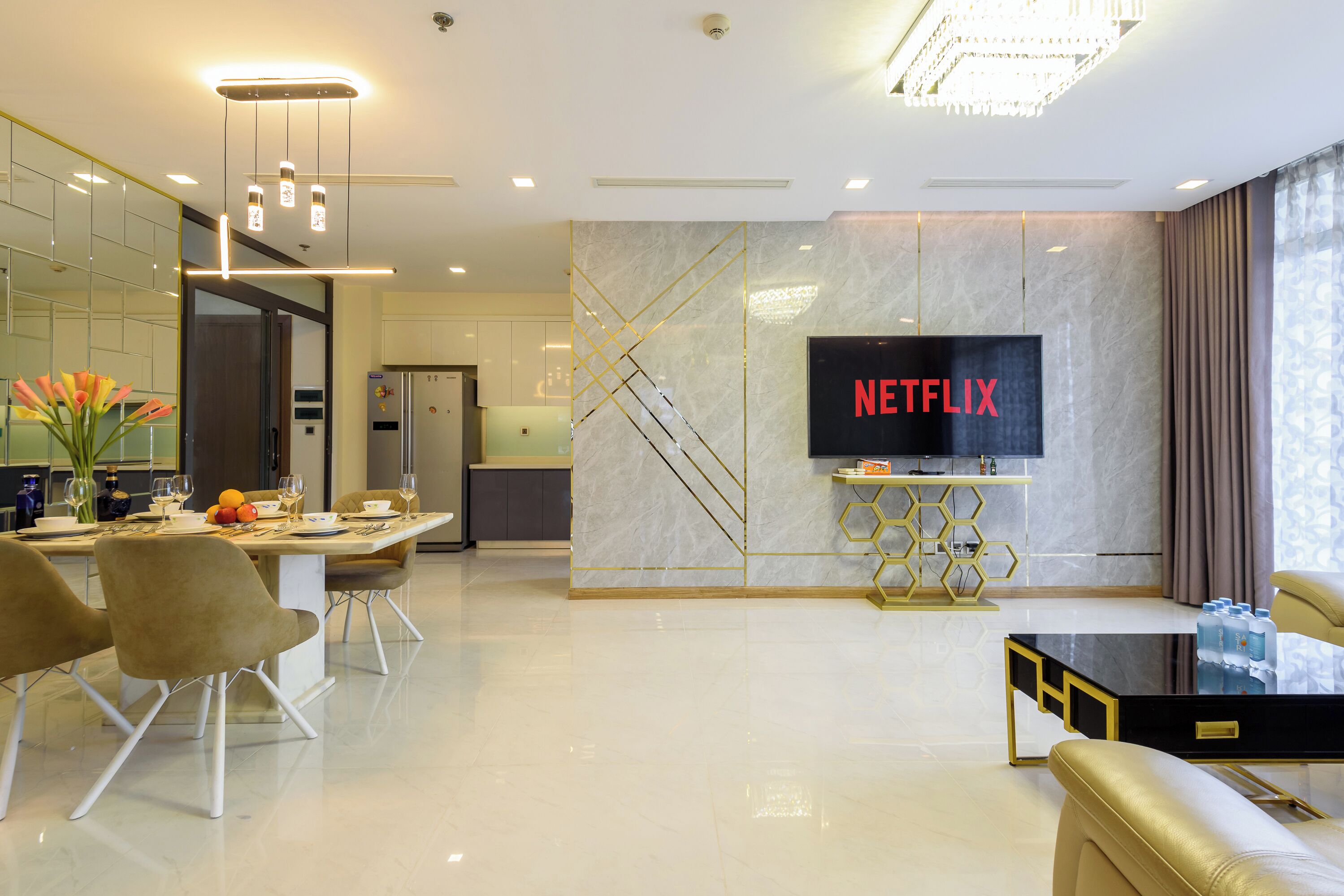 A modern living room with a large flat-screen TV, a dining area with a white table and chairs, and a kitchen in the background.