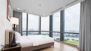 Habitación de hotel moderna con una cama grande, una mesita de noche con lámpara y vistas a un paisaje urbano a través de amplios ventanales.