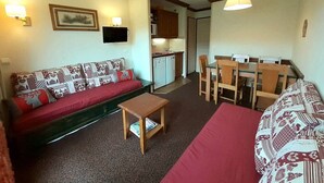 Un espacio habitable compacto con sofá, una mesita, zona de comedor con sillas, cocina con armarios y fregadero, y un televisor montado en la pared.