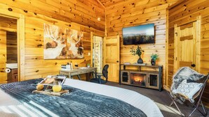 A cozy log cabin room with a bed, a desk, a fireplace, and a mounted TV.