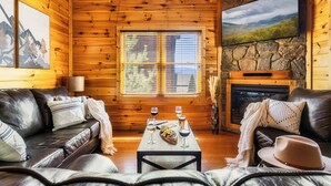 A cozy cabin interior with wooden walls, a stone fireplace, two leather sofas, a dining table with a meal and drinks, and a flat-screen TV mounted on the wall.