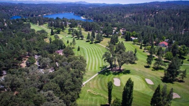 Vue aérienne d’un terrain de golf avec un lac, entouré de forêts denses et de zones résidentielles.