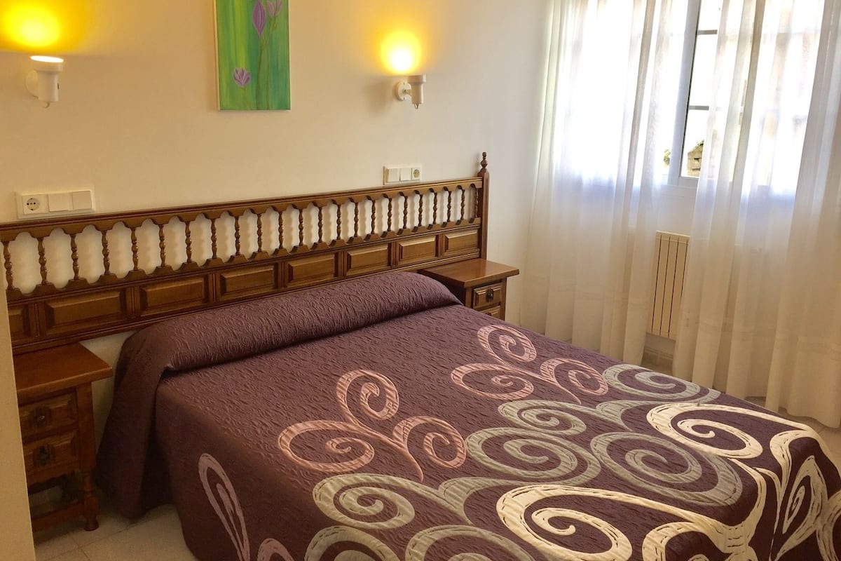 Un dormitorio con cabecera de madera, cama con cubrecama estampado, dos lámparas de pared y una ventana con cortinas traslúcidas.