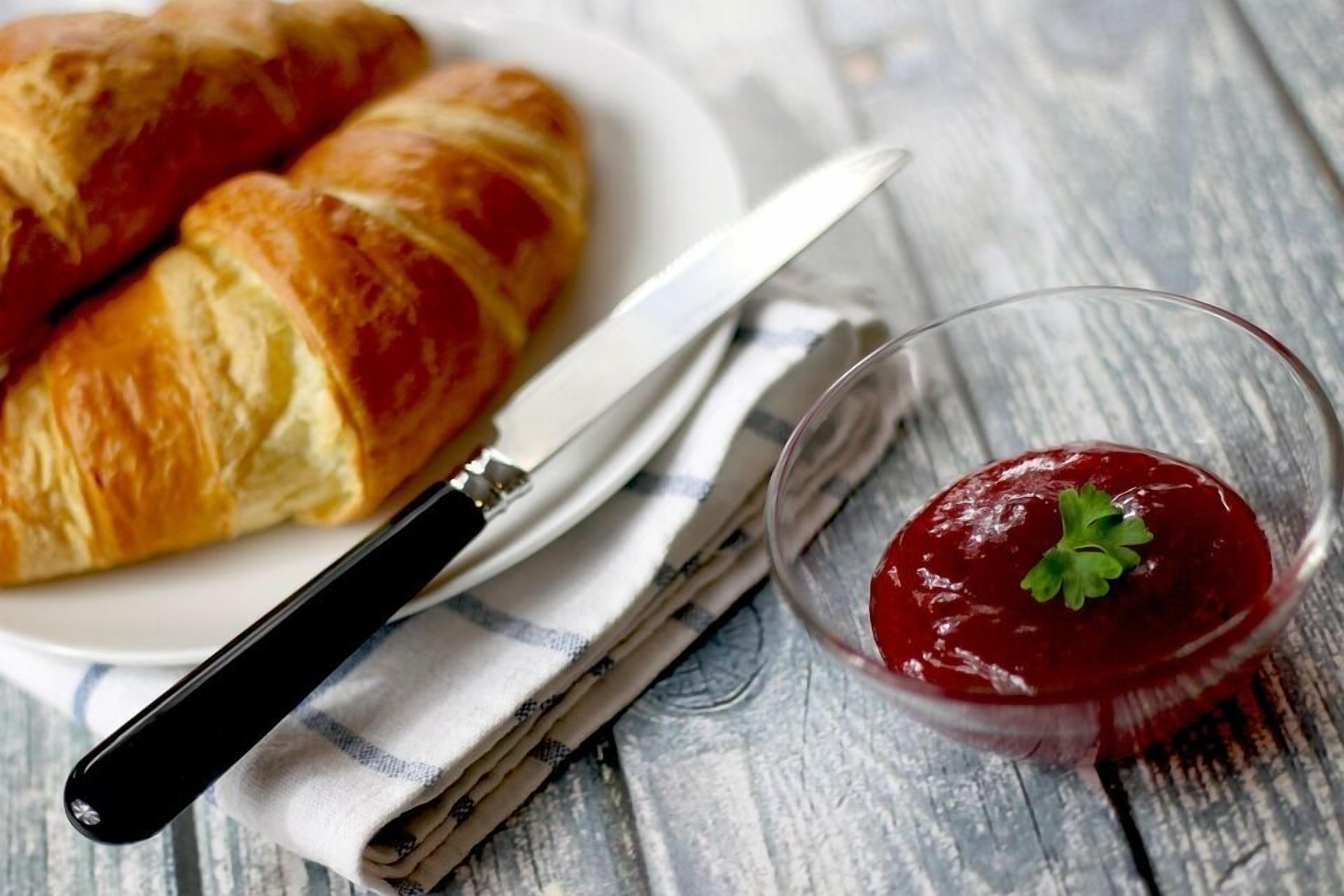 A croissant on a plate, a knife, and a glass of jam with a garnish.