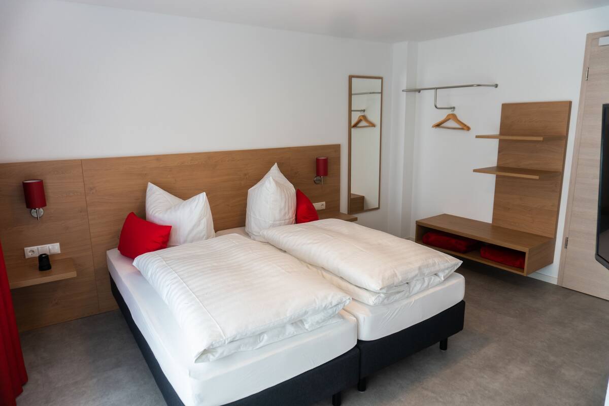 Cama doble con ropa de cama blanca y almohadas rojas, cabecera de madera, mesita de noche con una lámpara roja, espejo y estante de madera con una prenda colgada.