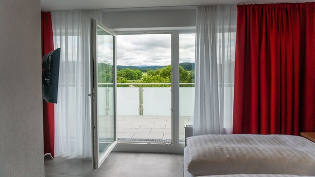 A hotel room with a bed, a TV, and a balcony with red curtains. - Pension Haas- Hotel am Turm (Rottweil)