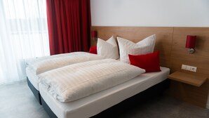 A double bed with white bedding and red pillows, a wooden headboard, a bedside table with a lamp, and a window with red curtains. - Pension Haas- Hotel am Turm (Rottweil)